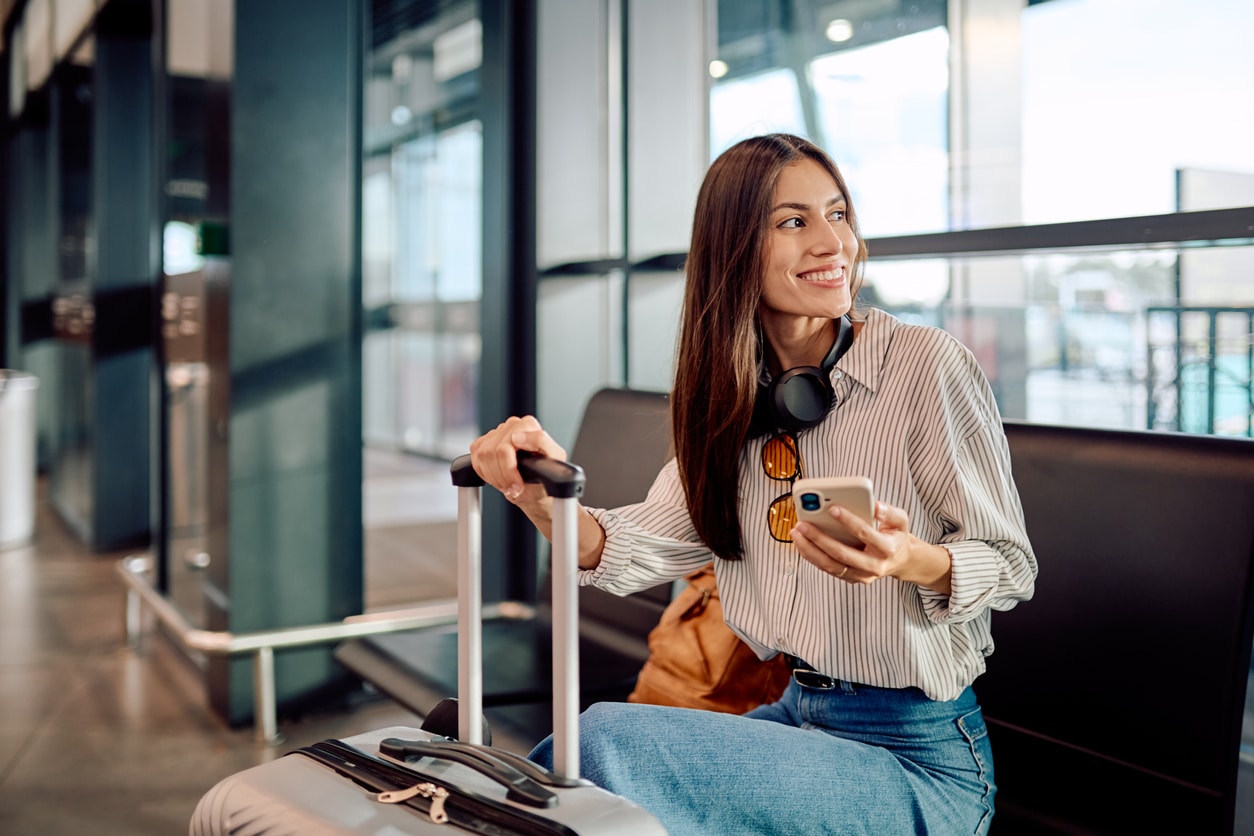 Smiling woman holding her trolley case and using her smartphone while waiting for her flight at the airport