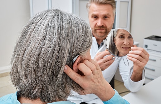 Senior woman trying on a hearing aid while looking into a mirror held by a hearing aid specialist in audiology clinic. Stylish hearing device.